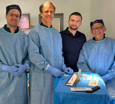 Group of four medical professionals in blue surgical gowns and gloves gathered around a prepared tray in an operating room setting.