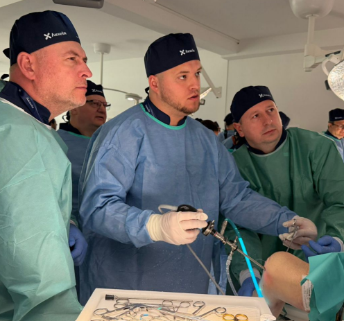 Surgeon in a blue gown uses surgical instruments during an operation, flanked by two assistants in green gowns in an operating room.