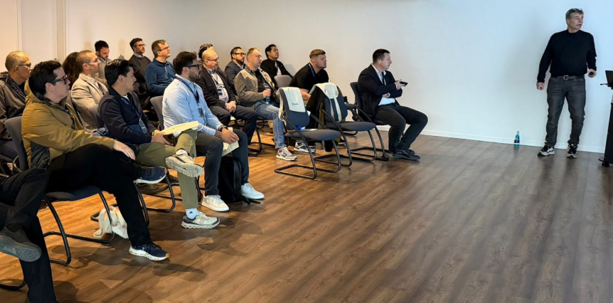 Group of professionals seated in a conference room listening to a speaker at the front of the room in casual attire.