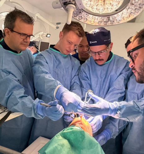 Surgeons in blue gowns perform an operation around a patient on an operating table under bright overhead lights.