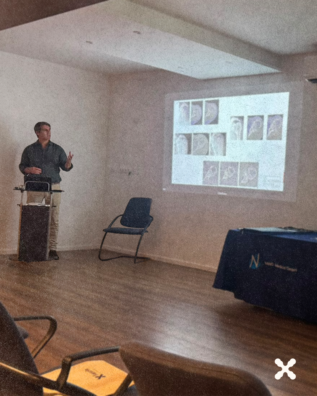Male presenter at a podium in a small lecture room, presenting a slide with a grid of portraits projected on the wall.