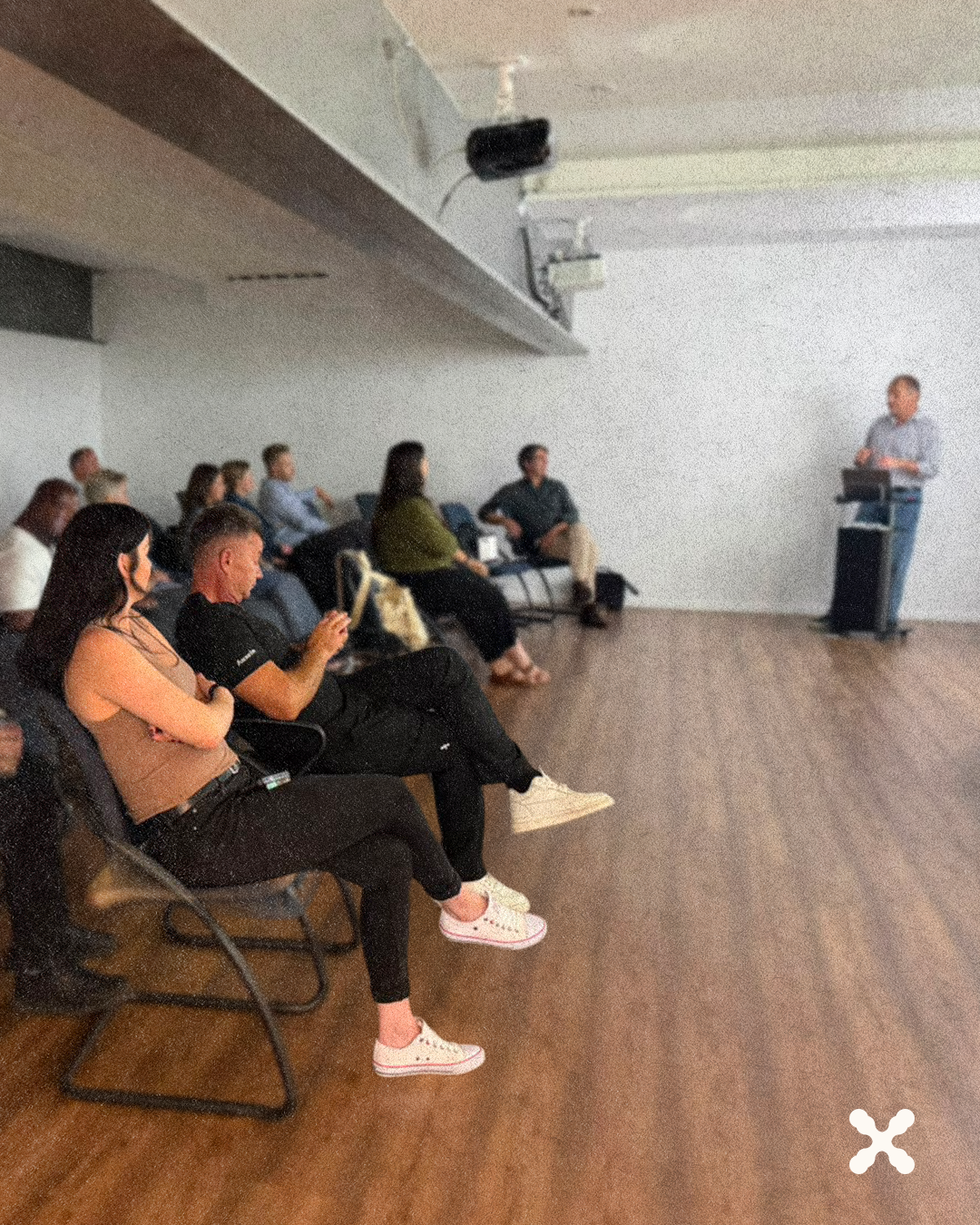 Presenter at a podium addressing a small audience in a meeting room with attendees seated along the wall.