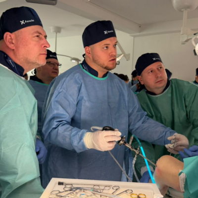 Surgeon in a blue gown uses surgical instruments during an operation, flanked by two assistants in green gowns in an operating room.