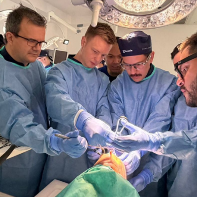 Surgeons in blue gowns perform an operation around a patient on an operating table under bright overhead lights.